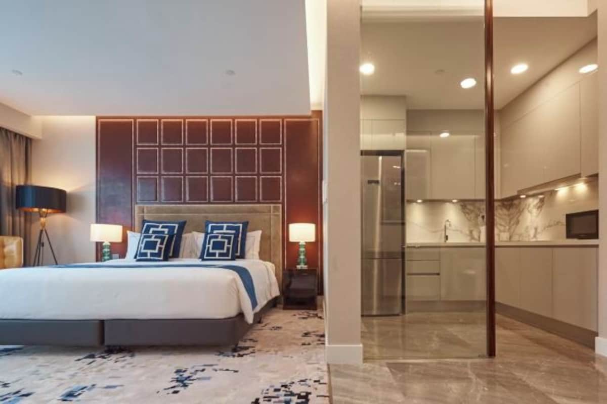 A modern hotel room with a large bed, bedside lamps, a headboard with a geometric pattern, a marble floor, and a view into the adjacent kitchen area.