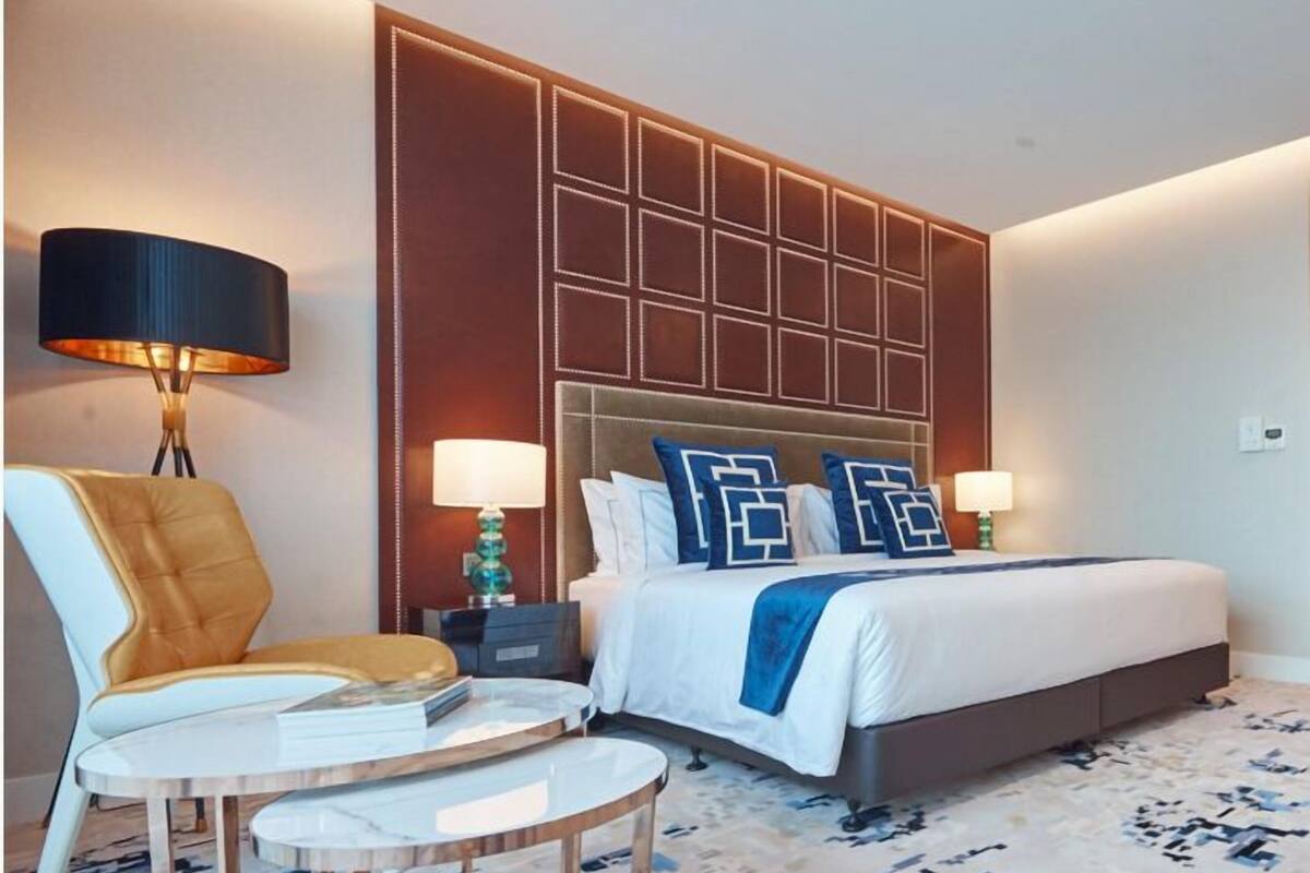 A hotel room with a large bed, two bedside lamps, a chair, a small table, and a decorative wall panel.