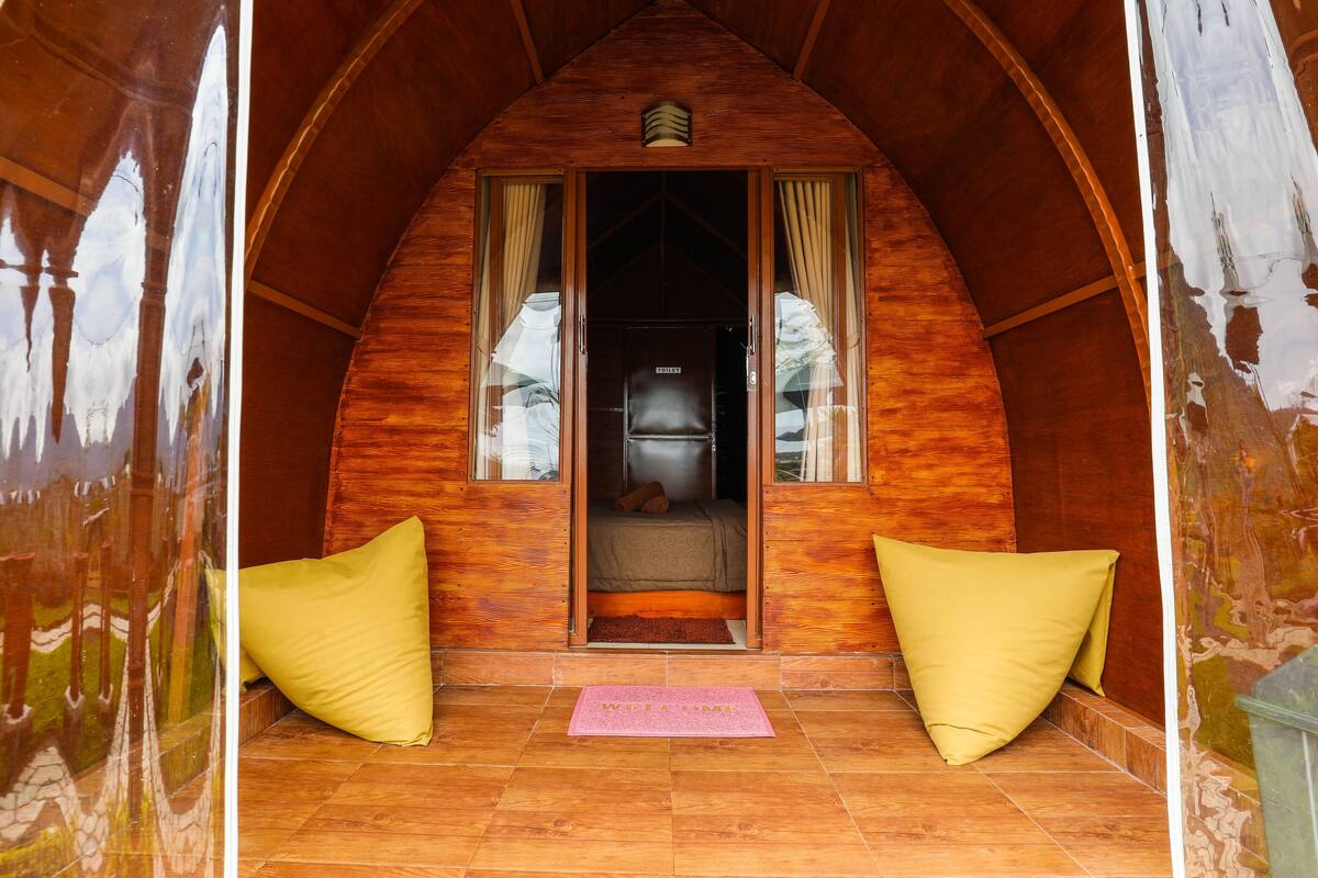 A cozy, wooden cabin-style room with a bed, a welcome mat, and two yellow pillows.
