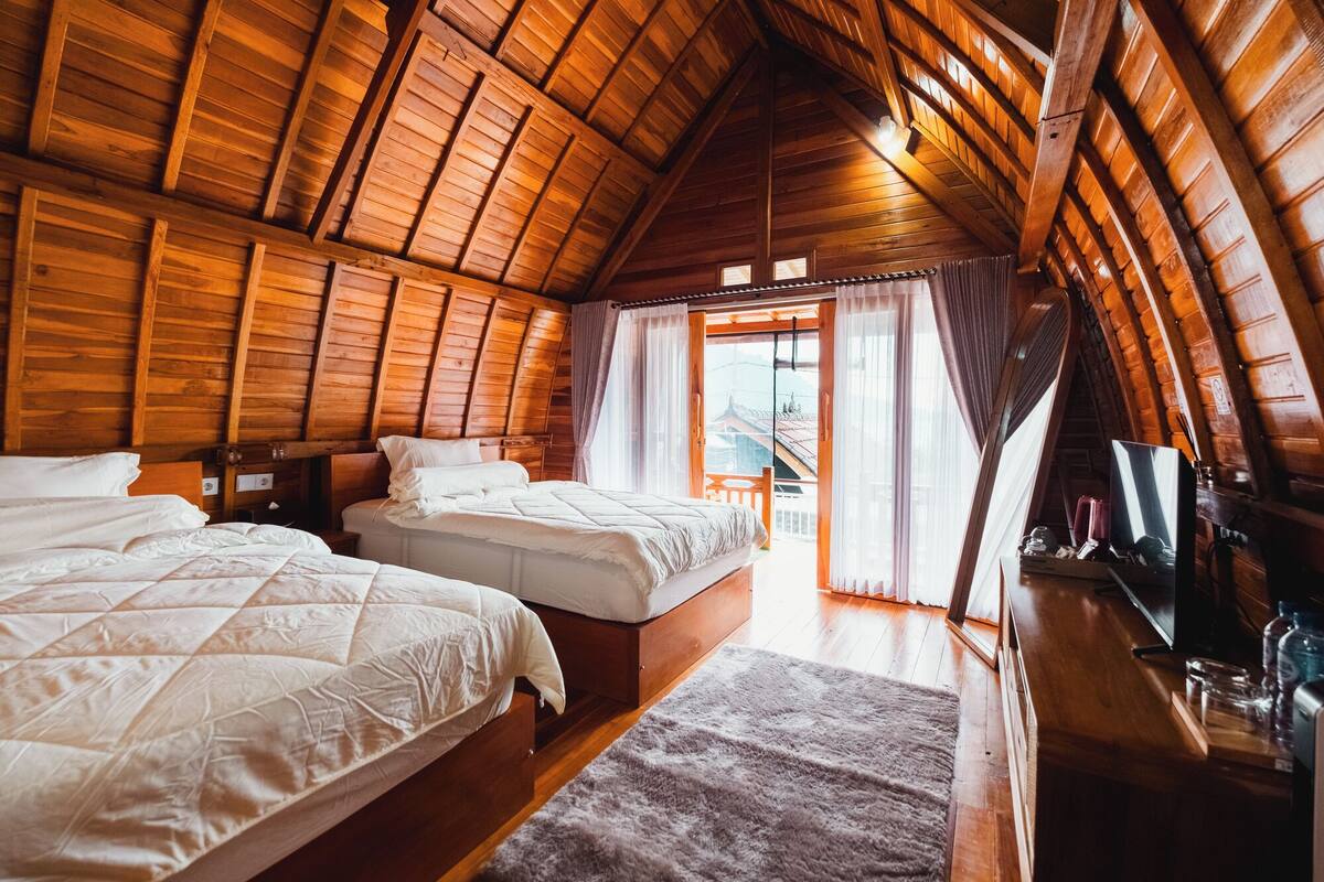 A cozy attic bedroom with two beds, wooden beams, and a view of a balcony.