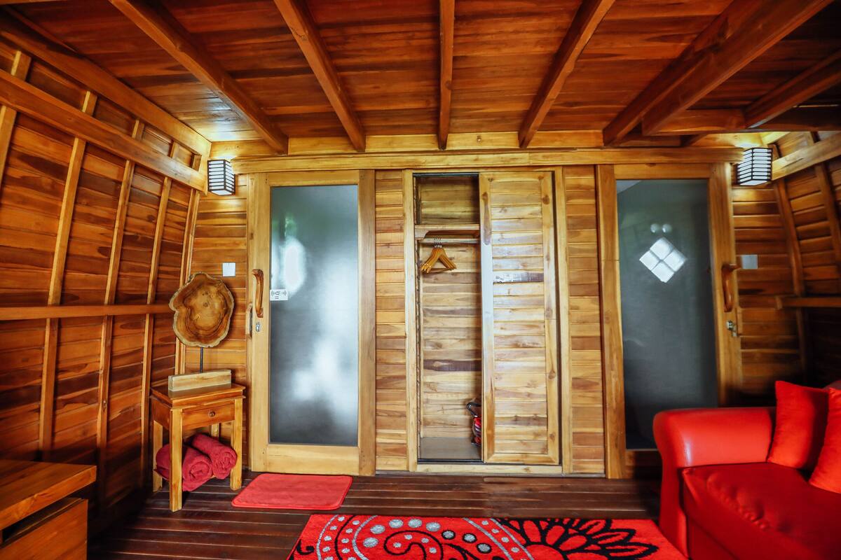 A wooden cabin interior with a sliding door, a red sofa, and a small wooden table with a hat and a red mat.