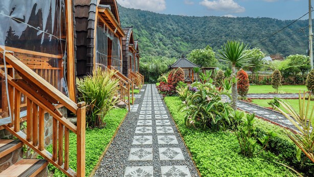 Una casa de madera con un sendero de baldosas que lleva a un quiosco tradicional, rodeado de exuberante vegetación y un paisaje montañoso.