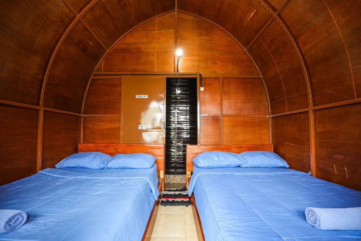 A room with two beds, a wooden ceiling, a door labeled "TOILET," and a small rug.