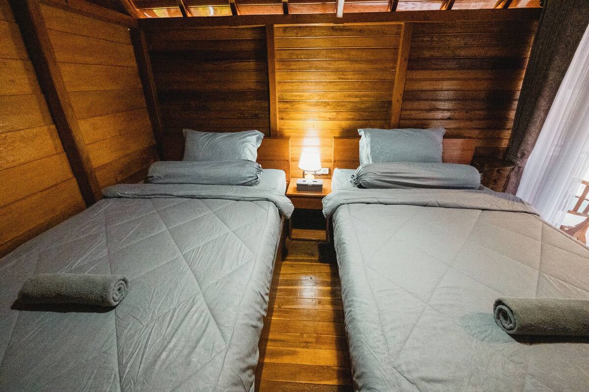 A small, cozy room with two single beds, wooden walls, and a nightstand with a lamp between the beds.