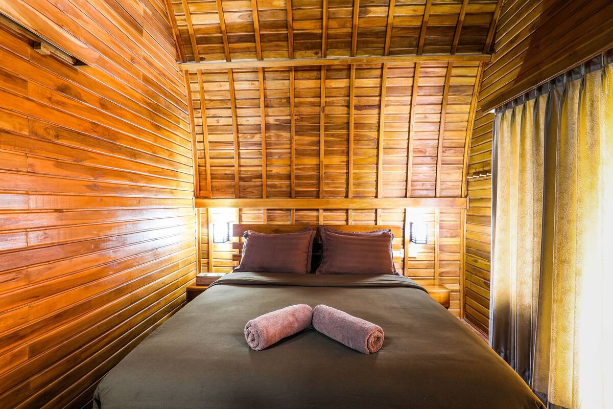 A wooden-paneled room with a bed, pillows, and a pair of rolled towels.