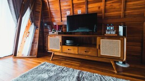Un interior de madera con un televisor sobre un mueble de madera, una ventana con cortinas y suelo de madera.