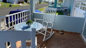 A balcony with a white rocking chair, a small table with a potted plant, and a view of a residential street with houses and palm trees. - Beach Getaway (Punta Santiago ,Humacao)