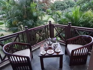 A balcony with a table set for tea, surrounded by wooden chairs, overlooking a lush garden. - Alaya resort Corbett (Ramnagar)