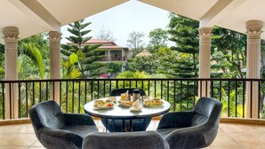 Un balcon avec une table ronde dressée pour le petit-déjeuner, entourée de deux chaises, donnant sur un jardin luxuriant et un bâtiment situé au loin.