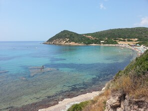 Une plage côtière aux eaux turquoise et limpides, bordée de rochers, avec des collines en arrière-plan.