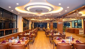 Een ruime eetzaal met meerdere tafels en stoelen, een bar in het midden en grote ramen langs de muren.