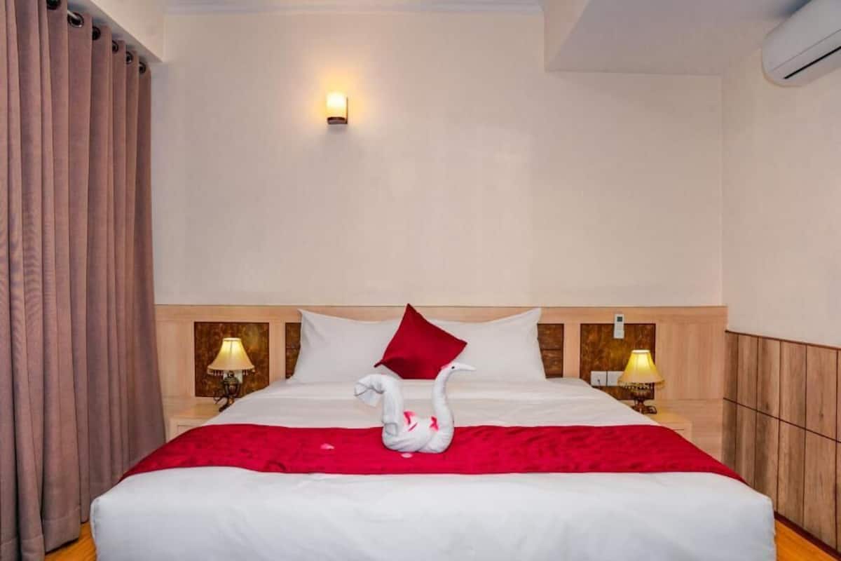 Een hotelkamer met een netjes opgemaakt bed, een rode plaid en een decoratief zwaan-kussen. Er staan twee nachtlampjes en er zit een lampje aan de muur.