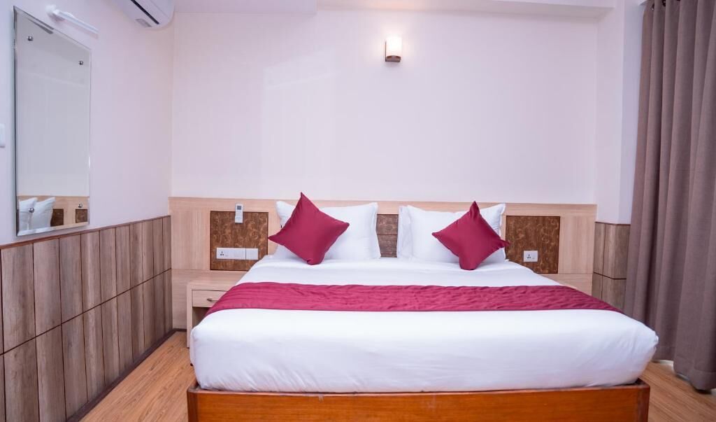 A hotel room with a wooden headboard, a large bed with white bedding and two red pillows, a nightstand, a mirror, and a wall-mounted light fixture.