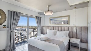 A modern hotel room with a large bed, a bedside table, a nightstand, a hanging light fixture, a view of the city through large windows, and a painting on the wall. - Knightsbridge E906 (Cape Town)