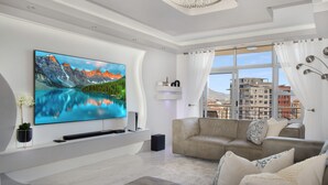 A modern living room with a large flat-screen TV, a beige sofa, and a view of buildings through large windows. - Knightsbridge E906 (Cape Town)