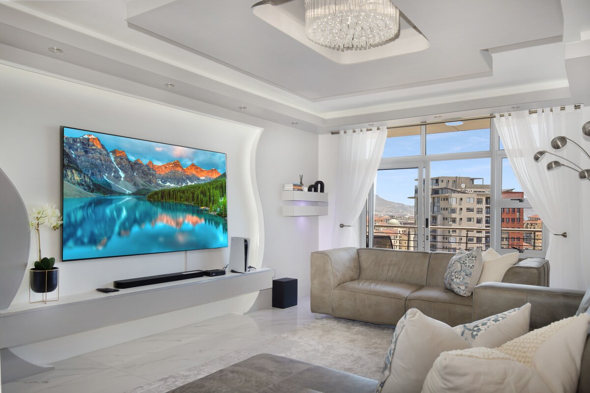 A modern living room with a large flat-screen TV, a beige sofa, and a view of buildings through large windows.