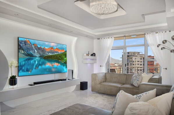 Una sala de estar moderna con un televisor de pantalla plana grande, un sofá color beige y vistas de edificios a través de ventanas amplias.