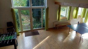 A room with a foosball table, a ping pong table, a bench, and large windows with curtains. - Vacation cottage Haus Hellhake (Sundern (Sauerland))