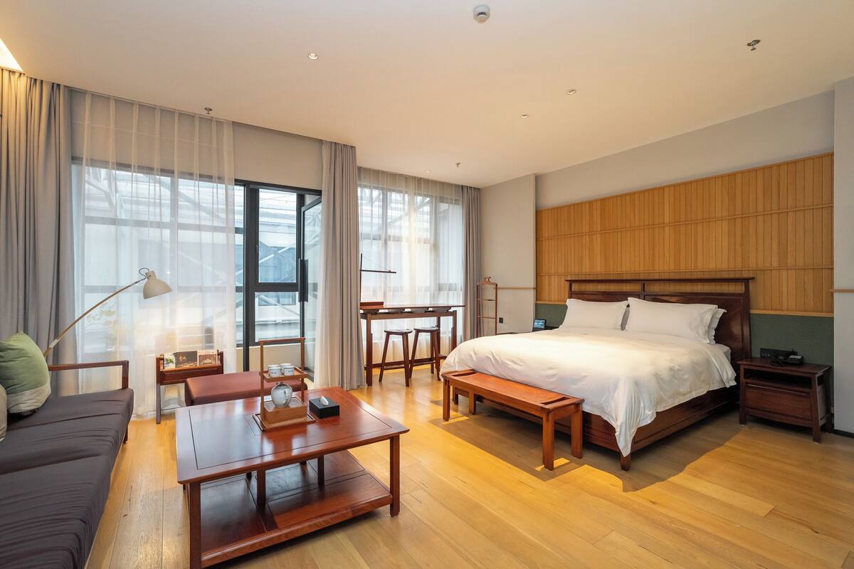 A modern hotel room with a large bed, a desk, a sofa, and a dining table.