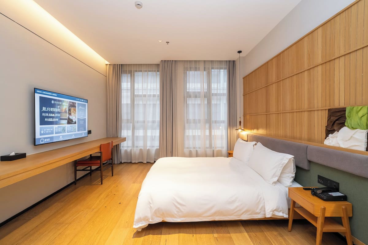 A hotel room with a bed, a desk, a chair, a TV mounted on the wall, and a window with curtains.