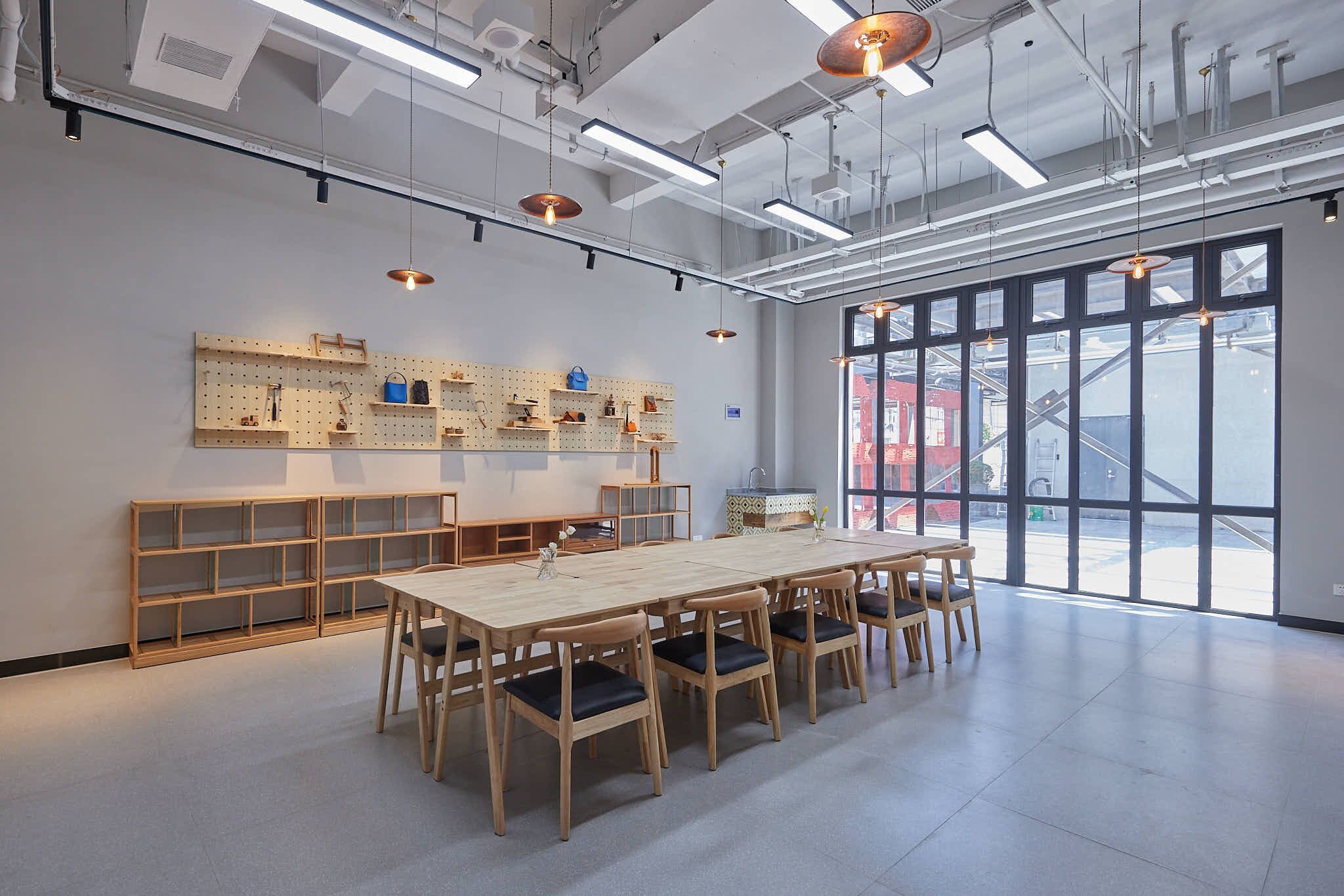 A spacious room with a long table and chairs, pegboards on the wall displaying various items, and large windows offering a view of the outside.