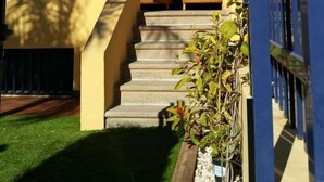 Ein zweistöckiges Gebäude mit gelber Fassade und blauen Akzenten, mit einer Treppe, die zu einer überdachten Veranda mit Sitzgelegenheit führt.