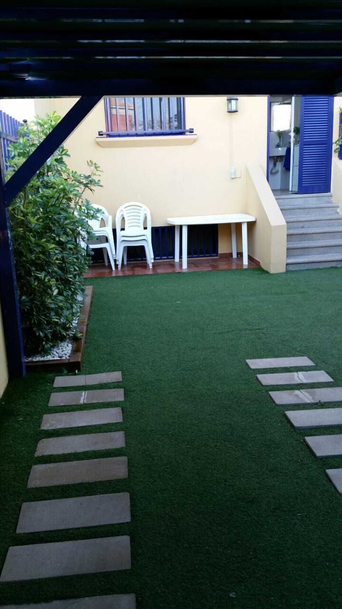 Een terras met kunstgras, witte plastic stoelen, een tafel en een trap die leidt naar een deur met blauwe luiken.