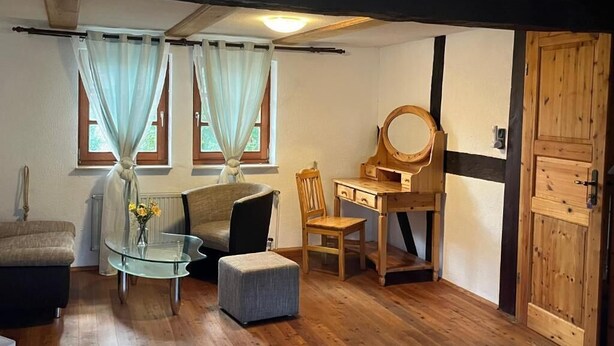 Una habitación con piso de madera, una mesa de vidrio con un jarrón de flores, un escritorio de madera con un espejo, una silla y un sofá.