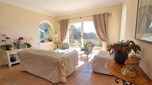 A sunlit room with a covered sofa, a small table with a potted plant, a chair, and a view through a window with curtains. - Air-conditioned luxury farmhouse, heated pool,sport hall, billiard table soccer, (Robion)
