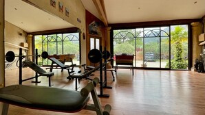 A gym with wooden flooring, exercise equipment, and large windows offering a view of greenery and a parked car. - Air-conditioned luxury farmhouse, heated pool,sport hall, billiard table soccer, (Robion)