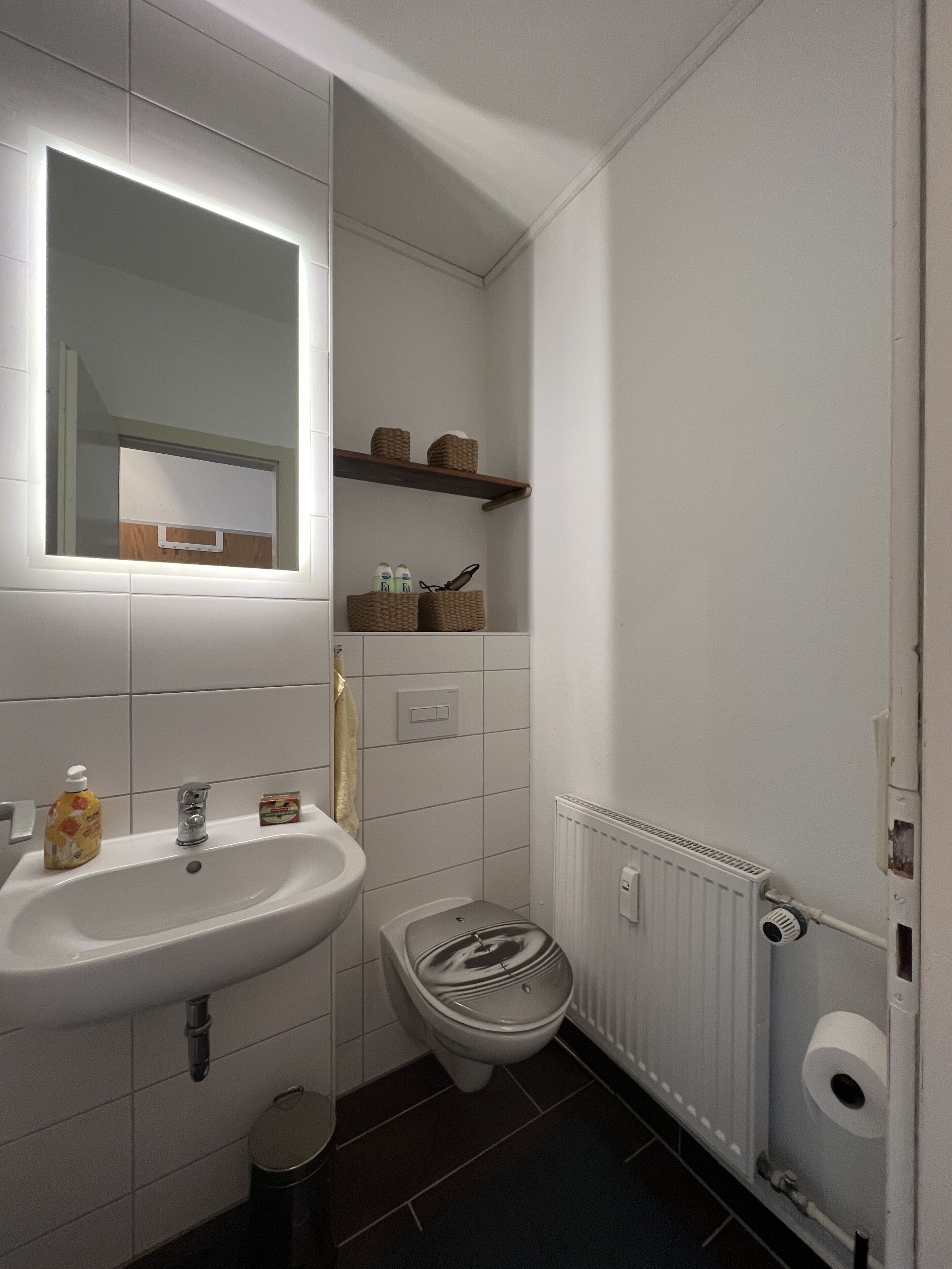 A bathroom with a white sink, mirror, and toilet. There are wicker baskets on a shelf above the toilet.