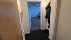 Een smalle gang met een houten deur links, een kapstok rechts en een deur aan het einde die leidt naar een kamer met een blauwe muur en een bed.