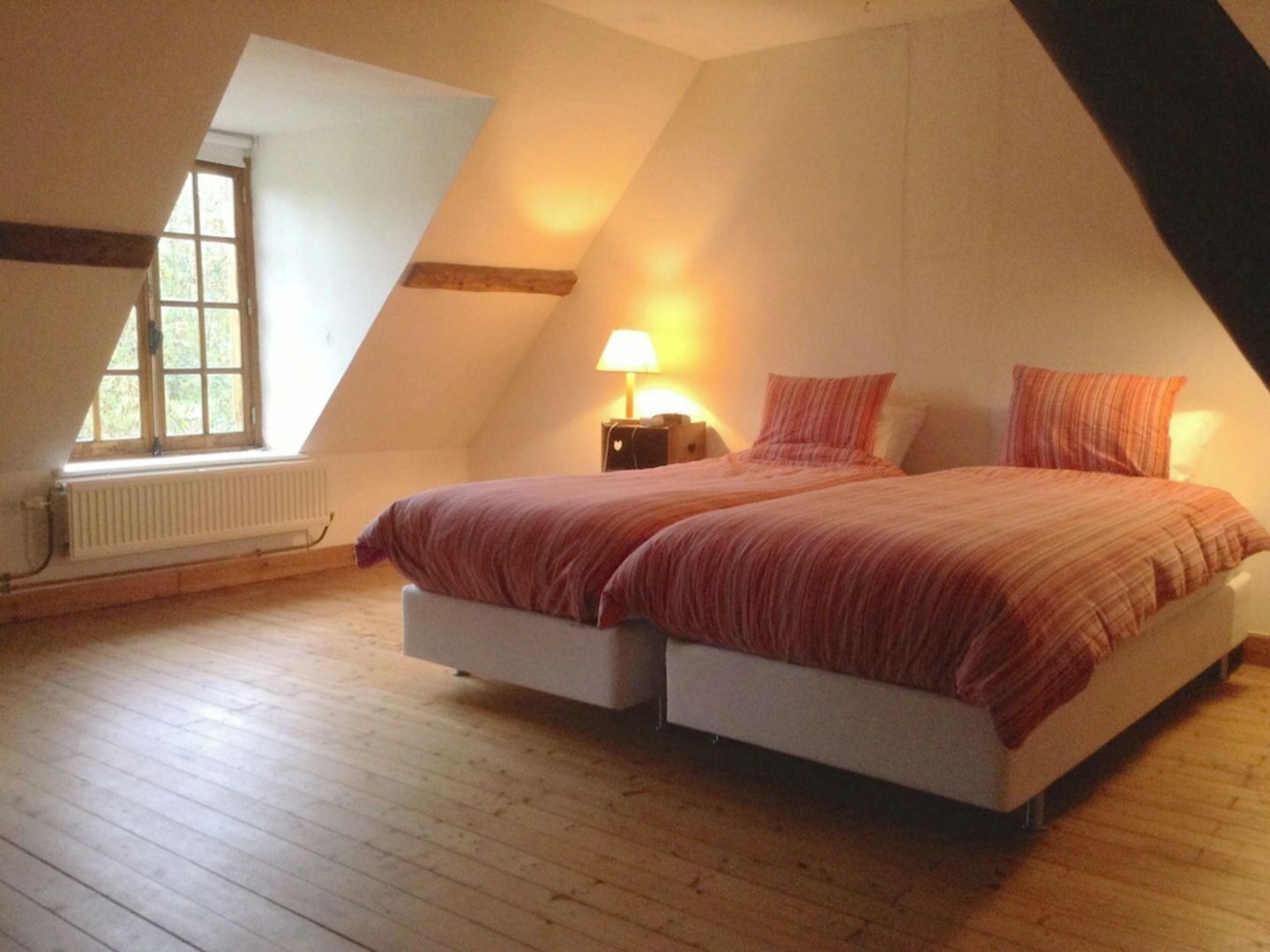 A bedroom with a sloped ceiling, a bed with two pillows, a nightstand, a lamp, and a window with a view of greenery.