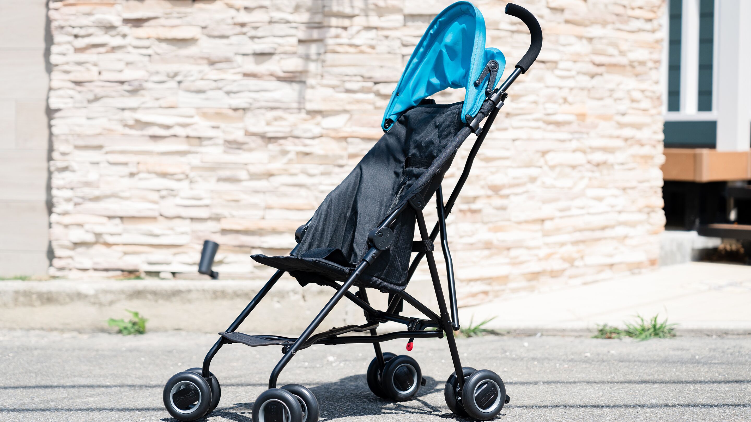 A black stroller with a blue canopy parked on a paved surface in front of a stone wall.