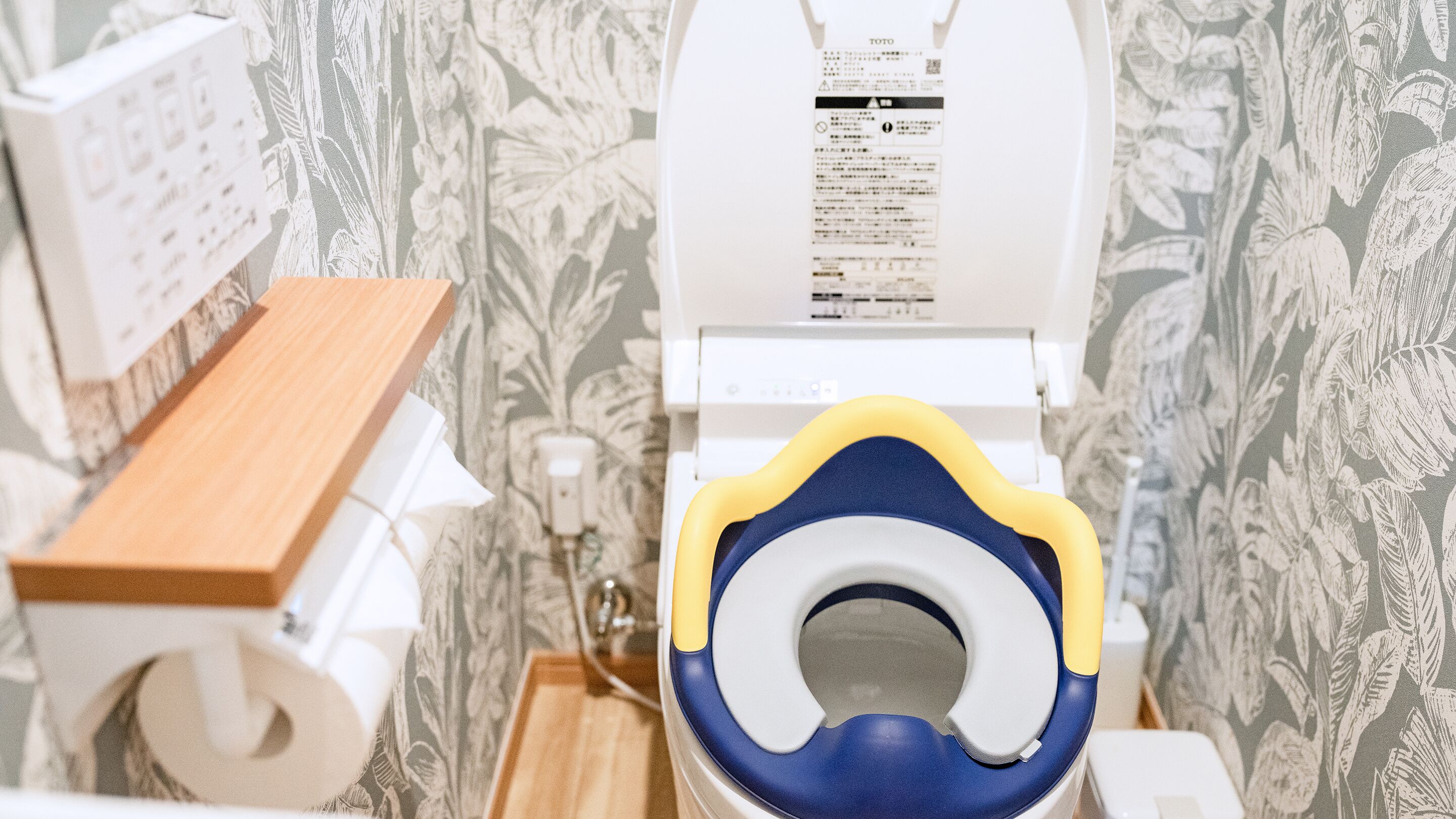 A bathroom with a toilet, a wooden shelf, and a wall with a floral pattern.