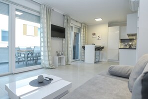 Un salon moderne avec un canapé, une table basse et un téléviseur. On aperçoit un coin repas avec des chaises et une table à travers une porte vitrée.