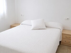 A neatly made bed with a white quilt, a single pillow, and a wooden nightstand with drawers.
