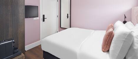 A hotel room with a bed, a TV mounted on the wall, a door, and a bedside table with a coffee machine.