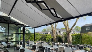 An outdoor cafe with tables and chairs, a glass enclosure, and a view of houses and trees. - Boutique Hotel Brasa Beemster (Middenbeemster)