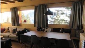 A cozy living room with a sofa, a dining table, and chairs. There are large windows with curtains, a lamp, and a view of trees. - Chalet Les Carlines (Courchevel)