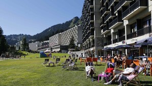 Et hotel med udendørs siddepladser, en græsplæne og et bjerglandskab i baggrunden.