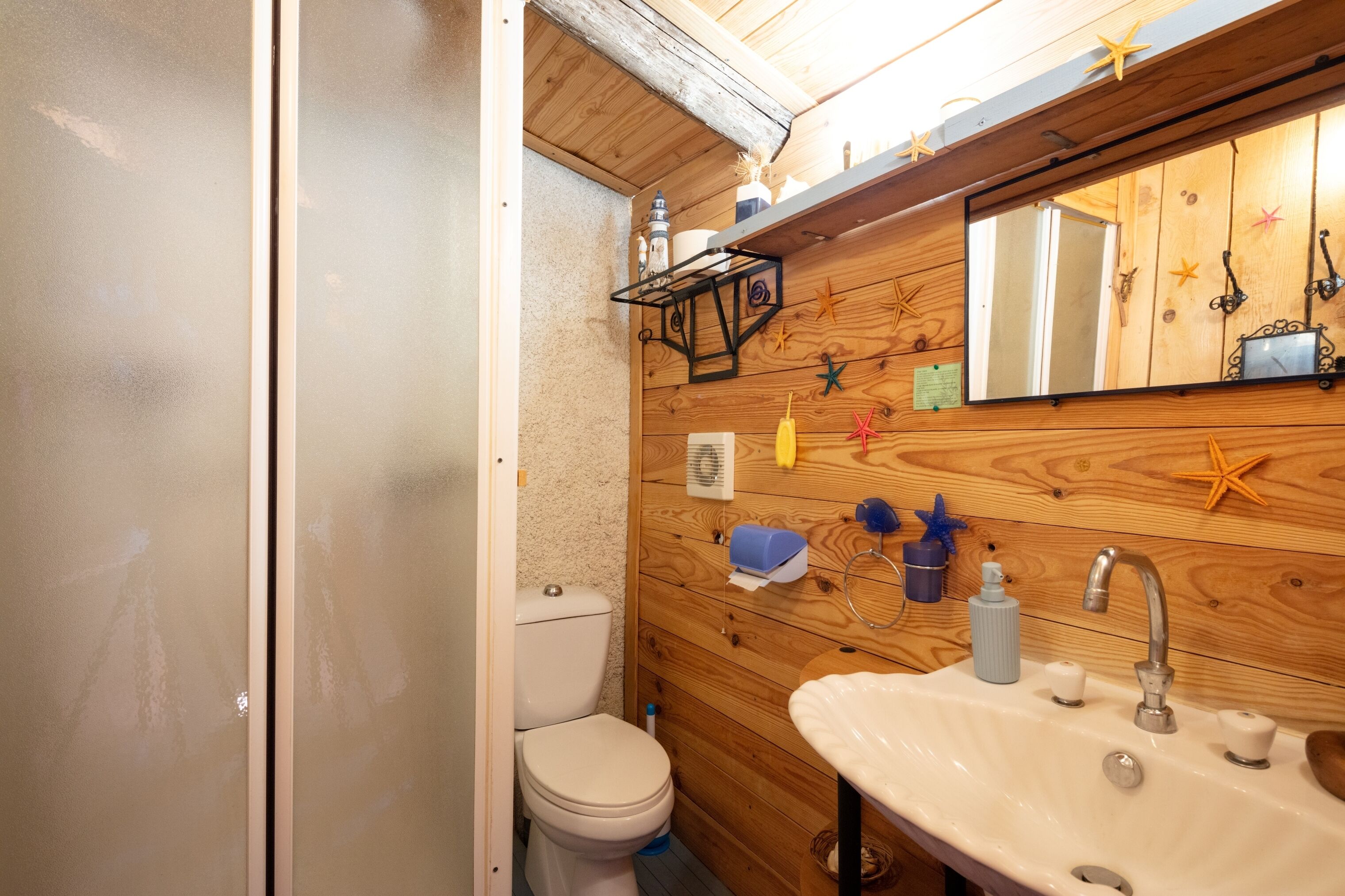 Une petite salle de bain avec un mur en bois, des toilettes blanches, un lavabo avec un miroir et divers produits de toilette.