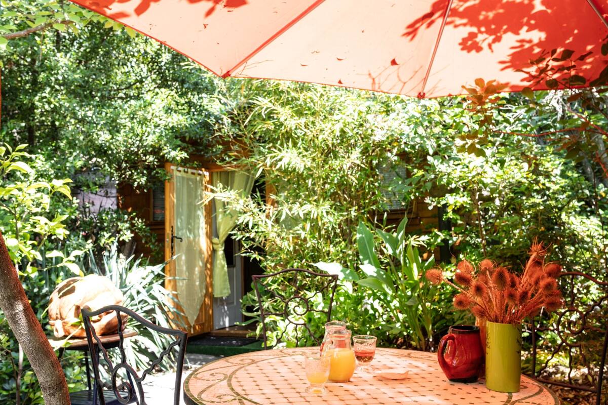 A round table with a red umbrella, a pitcher, a glass, and a vase with plants.