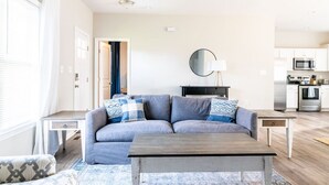 Ein modernes Wohnzimmer mit einem grauen Sofa, einem Holztisch und einem gemusterten Teppich. Im Hintergrund ist eine Küche mit weißen Schränken und Edelstahlgeräten zu sehen.