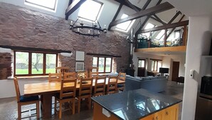 A spacious kitchen with a large island, wooden dining table, and chairs. There are multiple windows providing natural light and views of the outdoors. - Spacious Converted Barn on a Flower Farm, Devon Countryside, Family Friendly (Kentisbeare)