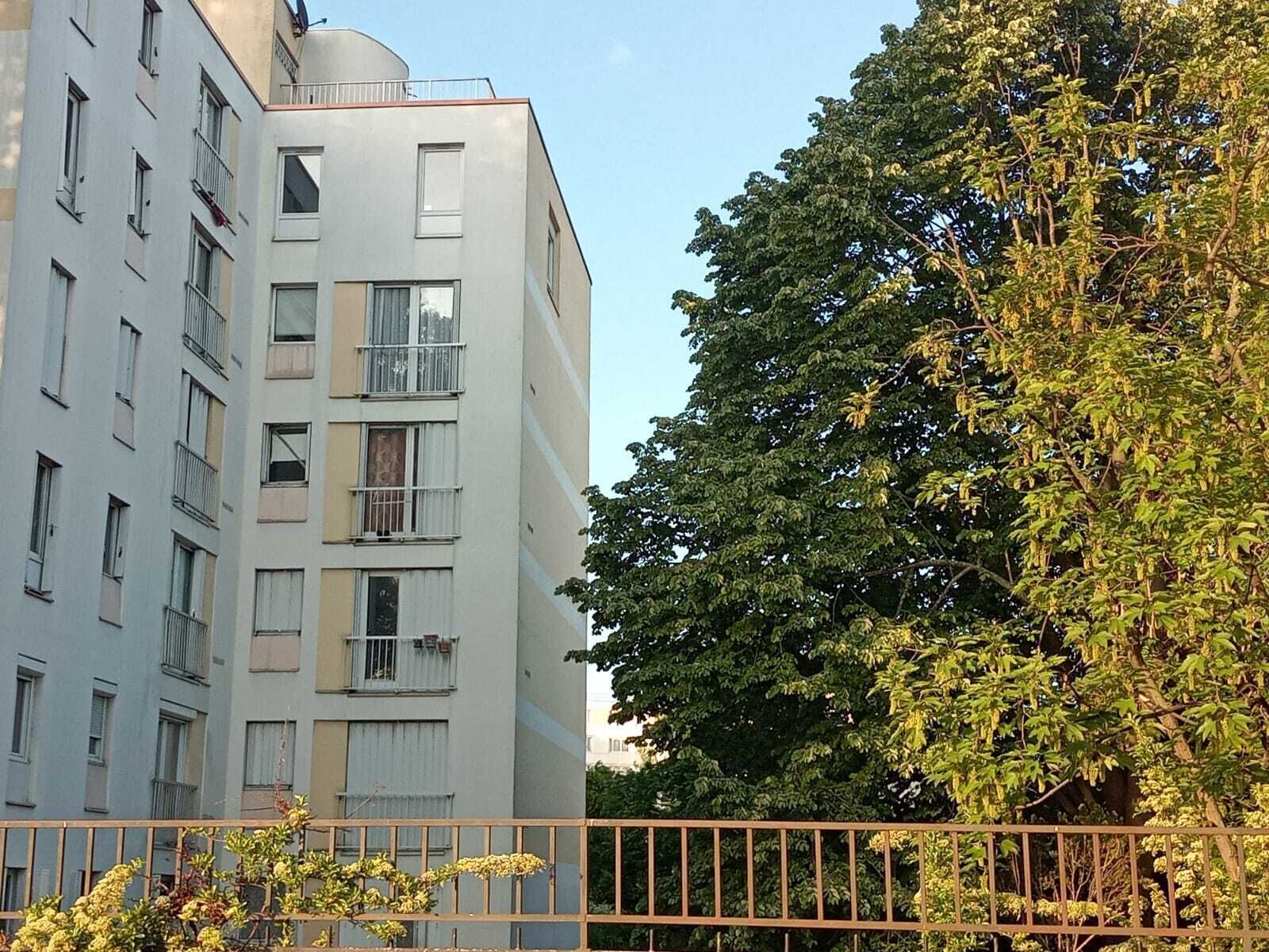 Un edificio de varios pisos con balcones, un árbol grande en primer plano y una cerca de metal.