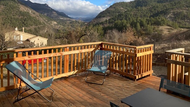 Una terraza de madera con dos sillas azules y una mesa, con vistas a un paisaje montañoso que incluye una casa al fondo.