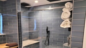 A modern shower area with a glass door, grey tiled walls, and a wooden bench. - Friary House Serviced Apartments by Roomsbooked - 24Hr Reception (Gloucester)