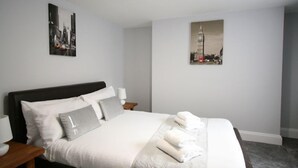A neatly made bed with pillows and a blanket, a bedside table with a lamp, and two framed pictures on the wall. - Friary House Serviced Apartments by Roomsbooked - 24Hr Reception (Gloucester)