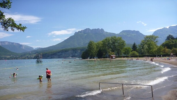 Ein See mit klarem Wasser, einem Sandstrand und Bergen im Hintergrund.
